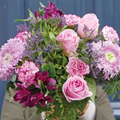Buquê de flores rosas, roxas e verde, segurado por pessoa com roupa bege em frente a parede azul