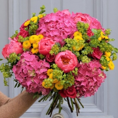 Buquê colorido de flores variadas com rosa, amarelo e verde, segurado por mão com manga castanha contra porta cinza.