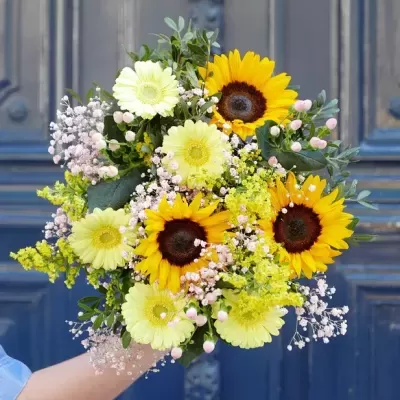 Ramo de flores com girassóis e flores pequenas coloridas segurado em frente a uma porta azul
