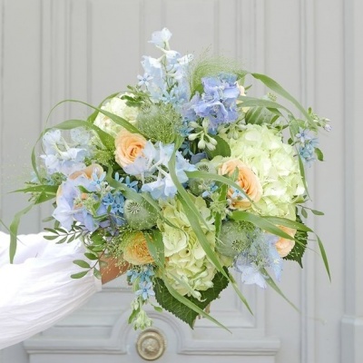 Buquê de flores com rosas pêssego, hortênsias verdes, flores azuis e folhagem contra porta cinza