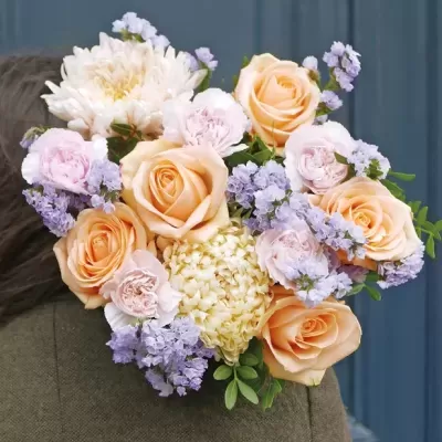 Bouquet de flores com rosas pêssego, peónias rosas e flores lilases sobre ombro de casaco cinzento
