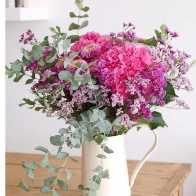 Arranjo floral rosa e lilás em jarra branca sobre mesa de madeira