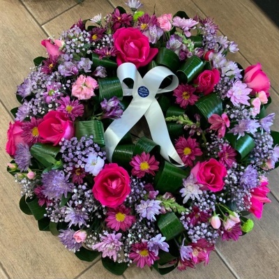 coroa de flores com rosas cor-de-rosa, flores roxas e brancas, folhas verdes, laço branco no centro