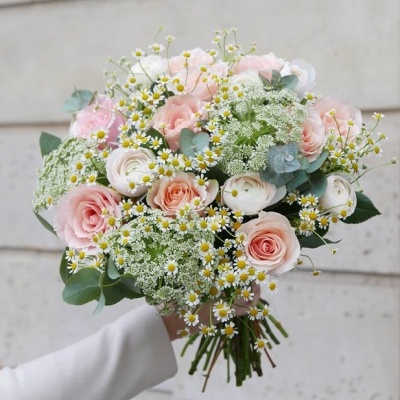 Ramo de flores com rosas cor-de-rosa, ranúnculos brancos e flores pequenas, segurado por uma mão com casaco branco
