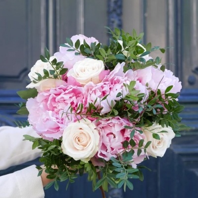 Bouquet de flores rosa e branca com folhagem verde segurado por mãos com mangas brancas em frente a uma porta azul