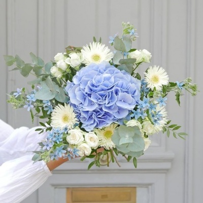 Buquê de flores azuis, brancas e verdes segurado por uma mão com manga branca.