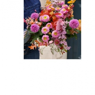 Ramo de flores rosadas, lilases e folhas alaranjadas nas mãos de uma pessoa com roupa azul escura