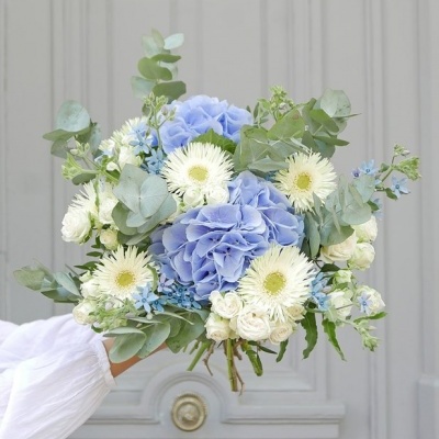 Bouquet de flores azuis e brancas com folhas verdes frente a uma porta cinzenta.