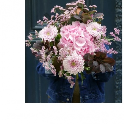 Buquê de flores cor-de-rosa com folhas verdes e pequenas flores, segurado por pessoa em casaco azul escuro