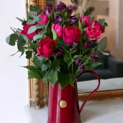 Jarro vermelho com flores rosas e roxas e folhas verdes sobre mármore junto a espelho dourado