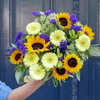 Bouquet colorido com girassóis, flores brancas e roxas e folhas verdes, contra uma porta azul