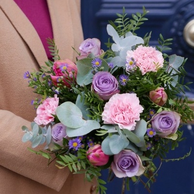 Buquê colorido com rosas, cravos e tulipas em tons de lilás e rosa com folhagem verde, segurado por pessoa com casaco bege e suéter bordô.