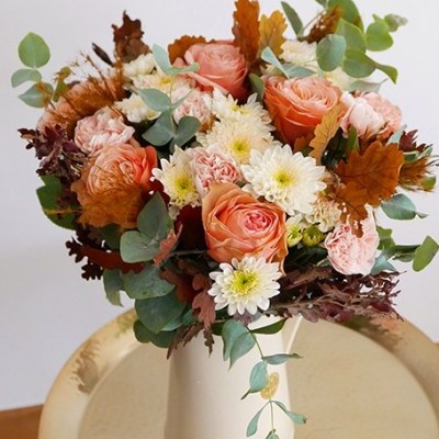 Arranjo floral com rosas, crisântemos e folhas em vaso branco sobre mesa dourada