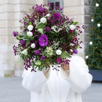 Bouquet grande de flores roxas e brancas com folhas verdes, segurado por pessoa com casaco branco em ambiente urbano