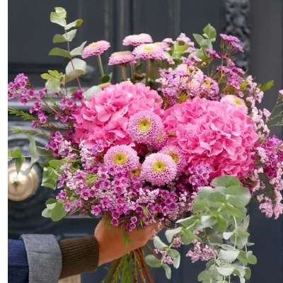 Buquê de flores cor-de-rosa com verdes segurado por mão em frente a porta preta.