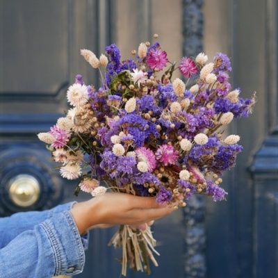 Ramo de flores secas roxas, brancas e rosas segurado por mão com casaco azul, em frente a porta azul escura