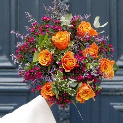Bouquet de flores com rosas laranja e flores pequenas, segurado à frente de porta azul