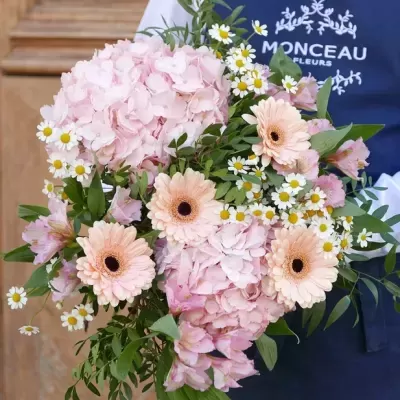 Buquê de flores cor de rosa com gerberas creme e pequenas flores brancas sendo segurado por pessoa com avental azul Monceau Fleurs