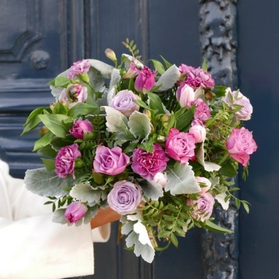 Bouquet de flores rosa e lilás com folhas verdes, segurado por pessoa com casaco branco diante de porta azul