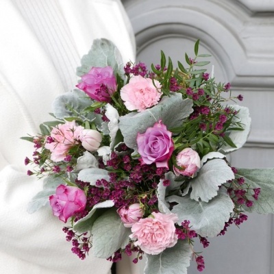 Buquê de flores rosa e lilás com folhagem prateada segurado por pessoa com casaco branco