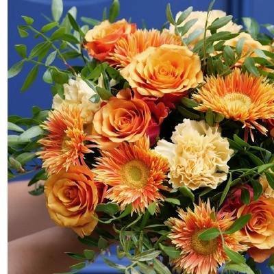 Bouquet de flores com rosas laranja, cravos creme e flores laranja e amarelas, folhagem verde, fundo azul