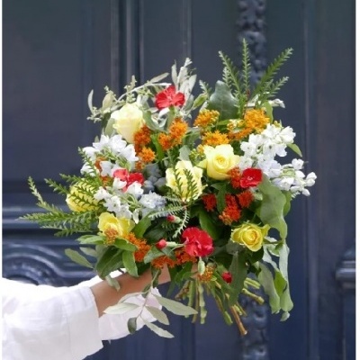 Buquê colorido de flores e folhas verdes segurado por mão com manga branca em frente a porta azul.