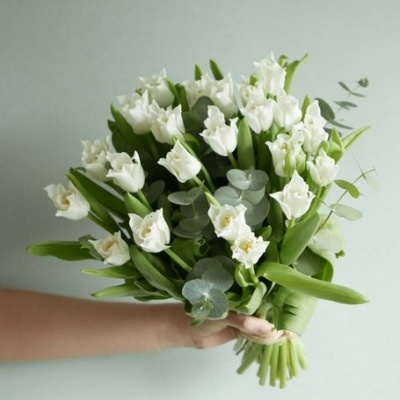 Bouquet de flores brancas com folhas verdes e ramos de eucalipto, mão segurando, fundo cinza claro