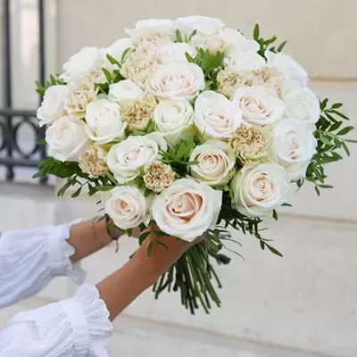 Buquê de rosas brancas com flores creme e folhagem verde, mãos em manga branca segurando