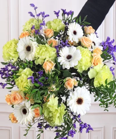 Buquê de flores variadas com margaridas brancas, rosas pêssego, flores verdes e ramos lilás seguidas por mão com manga preta.