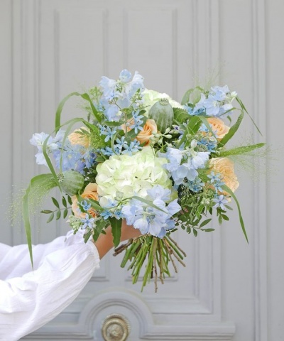 Buquê de flores azuis, brancas e pêssego com folhas verdes, segurado por uma mão com manga branca