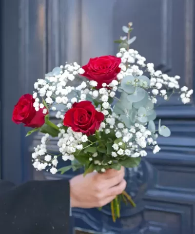 Buquê com rosas vermelhas, eucalipto e pequenas flores brancas segurado à frente de porta azul
