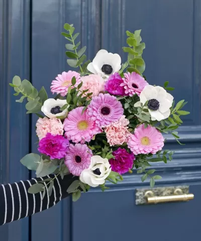 Ramo de flores com anêmonas brancas, cravos rosa, gerberas rosa e lilás, folhas verdes, contra porta azul