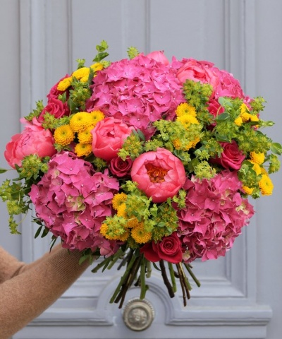 Buquê colorido de flores variadas com rosa, amarelo e verde, segurado por mão com manga castanha contra porta cinza.