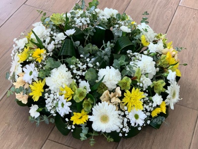 Arranjo floral artificial redondo com flores brancas, amarelas e verdes sobre chão de madeira.