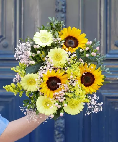 Ramo de flores com girassóis e flores pequenas coloridas segurado em frente a uma porta azul
