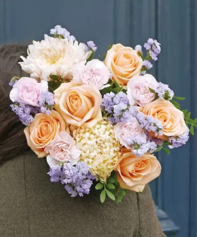 Bouquet de flores com rosas pêssego, peónias rosas e flores lilases sobre ombro de casaco cinzento