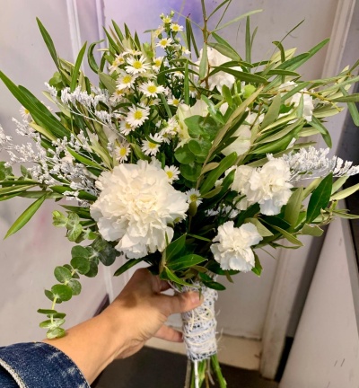 Ramo de flores brancas com folhagem verde amarrado com fita branca, segurado por uma mão