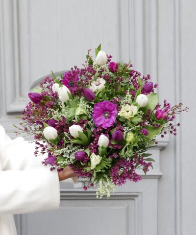 Ramo de flores roxas e brancas com folhagem diversificada, diante de uma porta cinzenta.