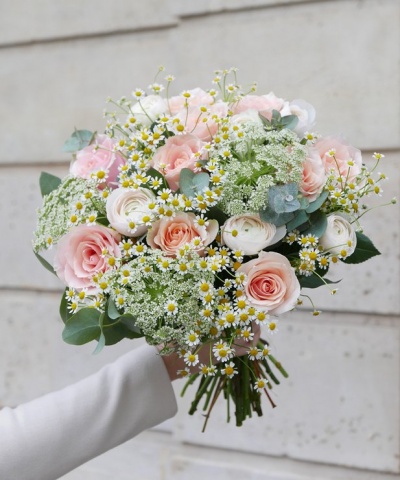 Ramo de flores com rosas cor-de-rosa, ranúnculos brancos e flores pequenas, segurado por uma mão com casaco branco