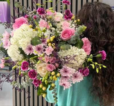 Ramo colorido de flores variadas com rosas, cravos, hortênsias e outras plantas, segurado por pessoa de cabelo encaracolado