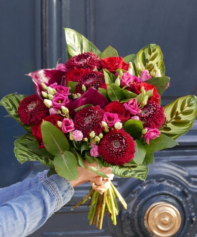 Buquê de flores vermelhas e rosas com folhas verdes seguras por uma mão diante de uma porta escura com detalhe dourado