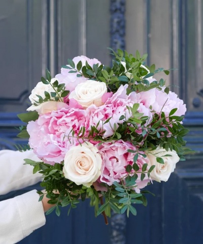 Bouquet de flores rosa e branca com folhagem verde segurado por mãos com mangas brancas em frente a uma porta azul