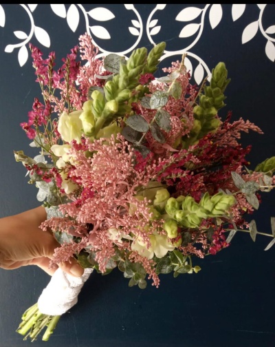 Buquê de flores verdes, rosas e brancas com folhagem, segurado por mão contra fundo azul escuro com padrão branco