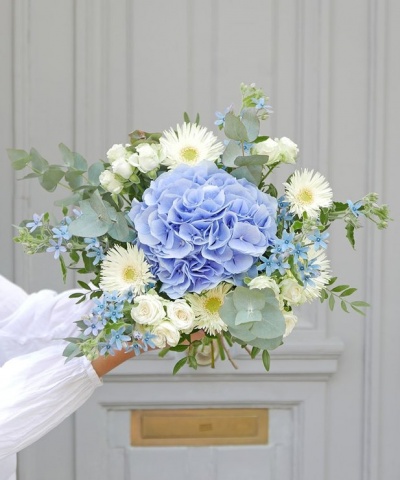 Buquê de flores azuis, brancas e verdes segurado por uma mão com manga branca.