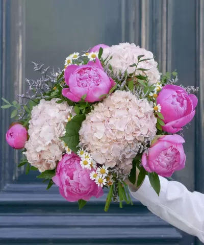 Bouquet com peónias rosa, hortênsias brancas e margaridas pequenas com folhagem, segurado em frente a porta azul