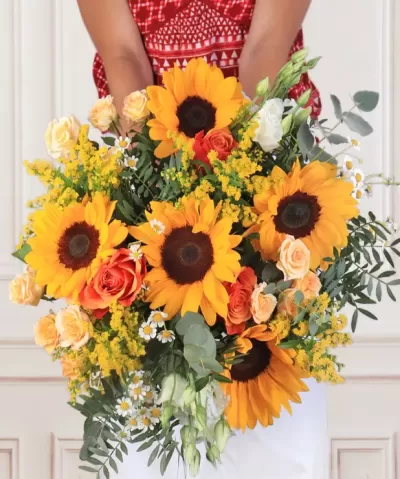 Arranjo floral com girassóis, rosas e folhagem segurado por pessoa com vestido vermelho