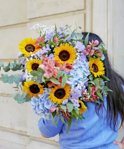 Arranjo colorido de flores com girassóis sendo segurado por pessoa de camisa azul