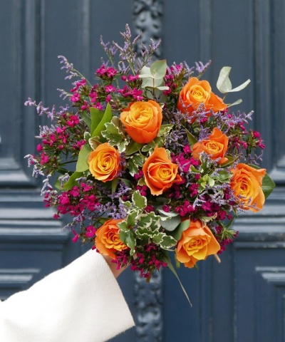Bouquet de flores com rosas laranja e flores pequenas, segurado à frente de porta azul