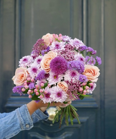Ramo de flores com rosas e crisântemos segurado por mão com camisa de ganga diante de porta escura