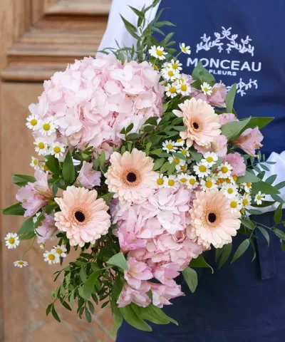 Buquê de flores cor de rosa com gerberas creme e pequenas flores brancas sendo segurado por pessoa com avental azul Monceau Fleurs
